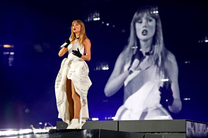 Singer performing on stage in a stylish dress with a high slit, holding a microphone. Large screen in background shows her image