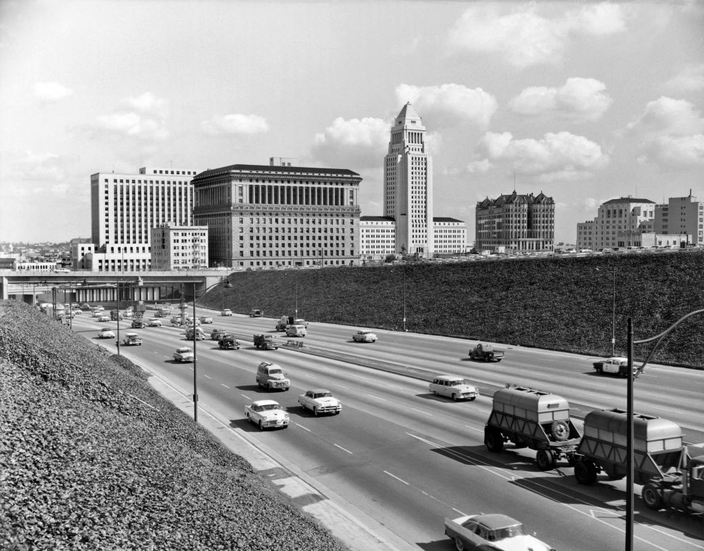 Historic photo of mid-20th century Los Angeles with cars on a highway and downtown buildings, including a notable tower in the background