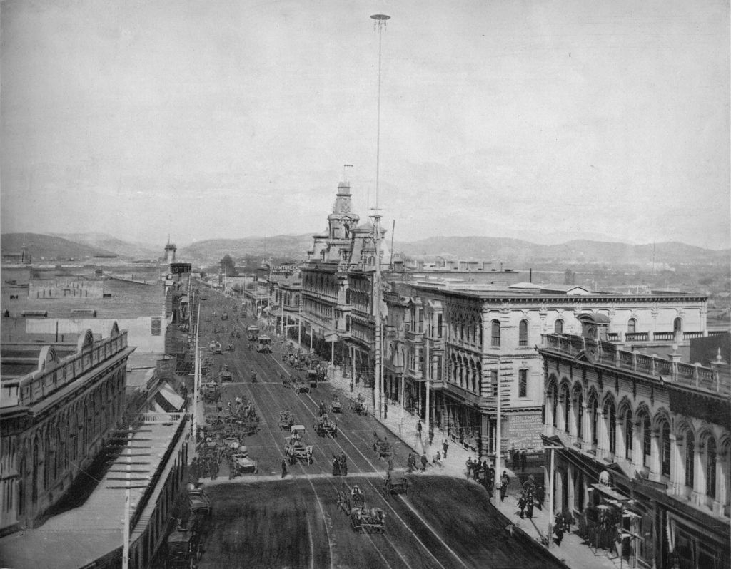 Historic black-and-white photo depicting a bustling city street with horse-drawn carriages and tall buildings lining both sides