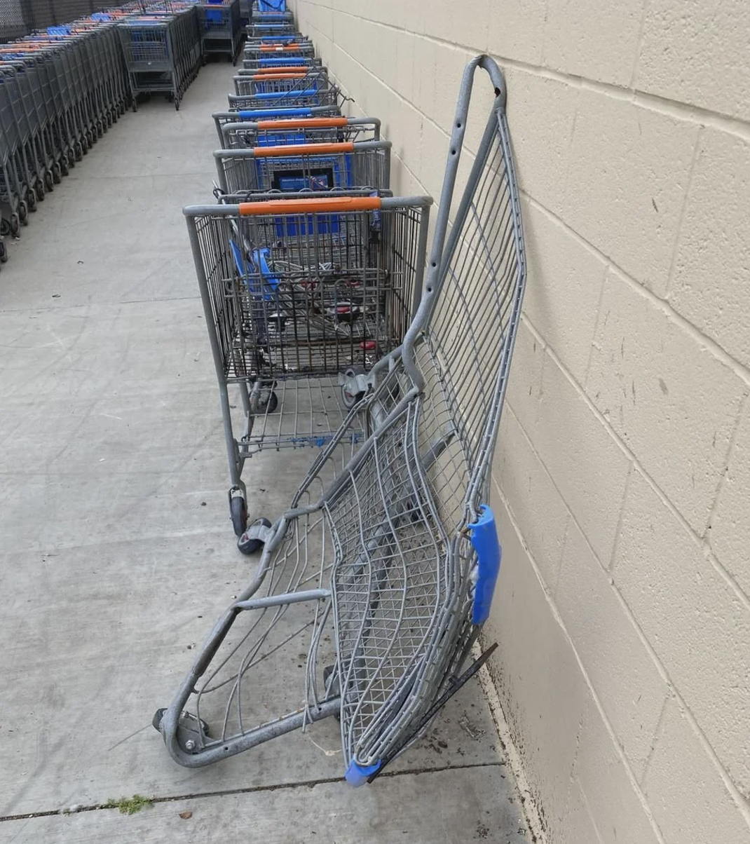 Bent shopping cart leans against a wall in a parking lot, with several intact carts lined up behind it