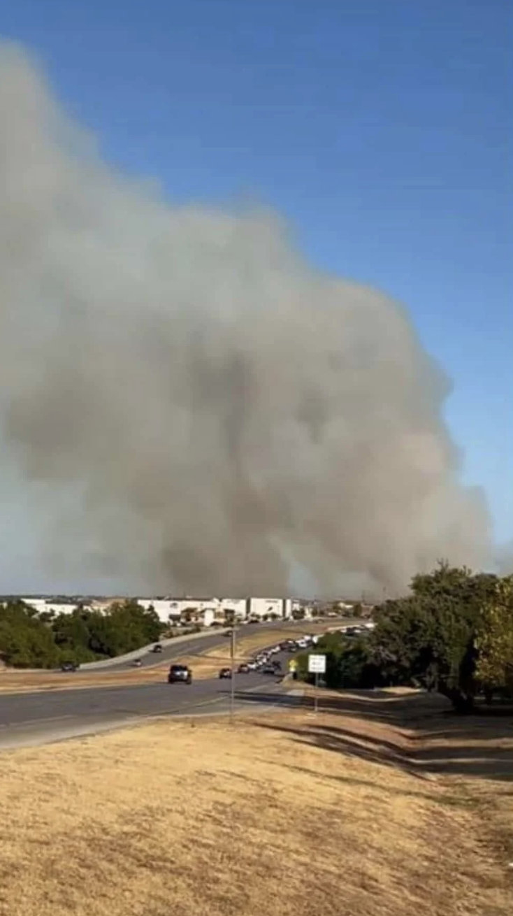 Large smoke plume rises over distant buildings and trees near a road with scattered cars, indicating a possible fire