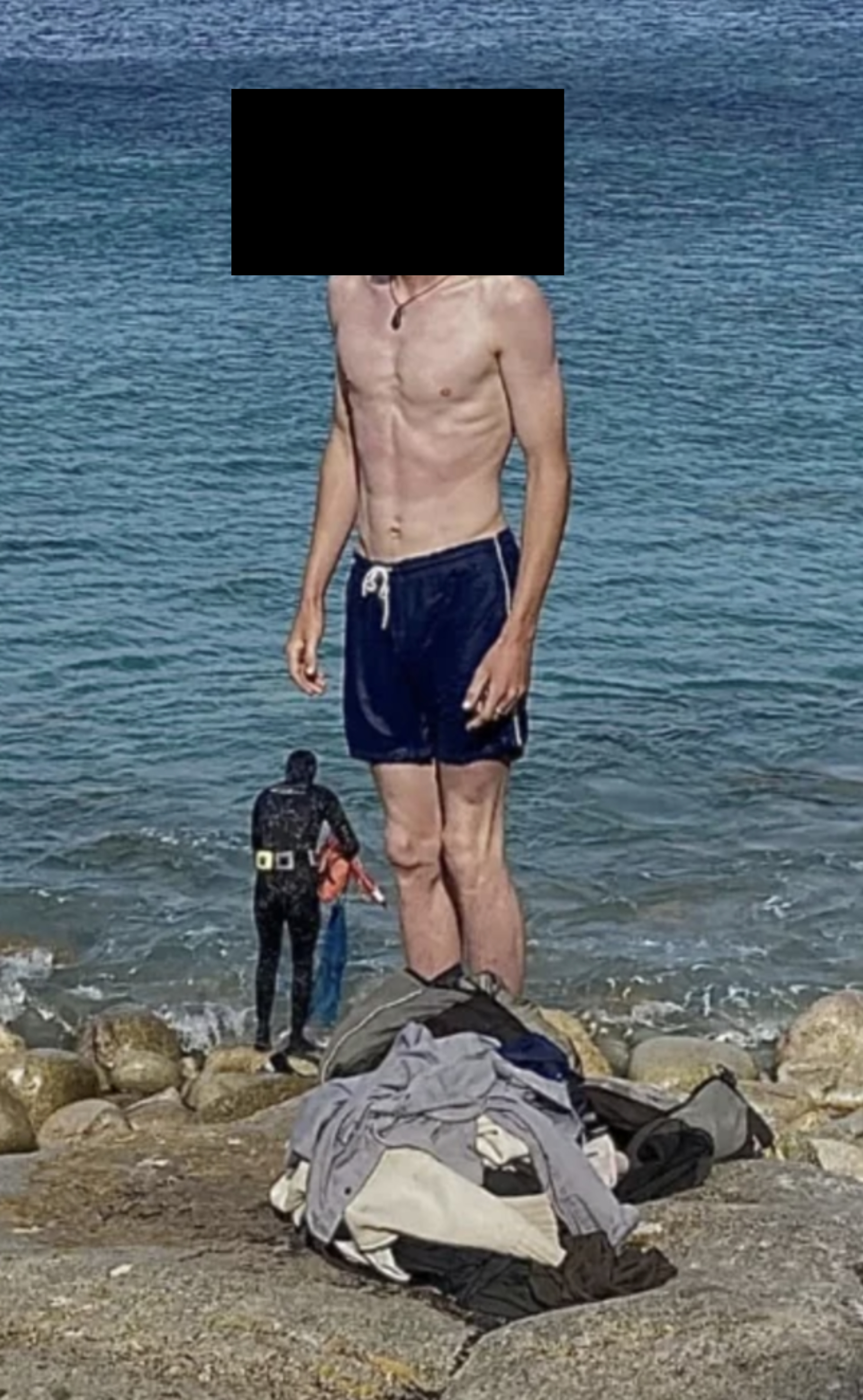 Person standing on rocks at a beach, wearing swim shorts. Waves and two people in wetsuits visible behind them