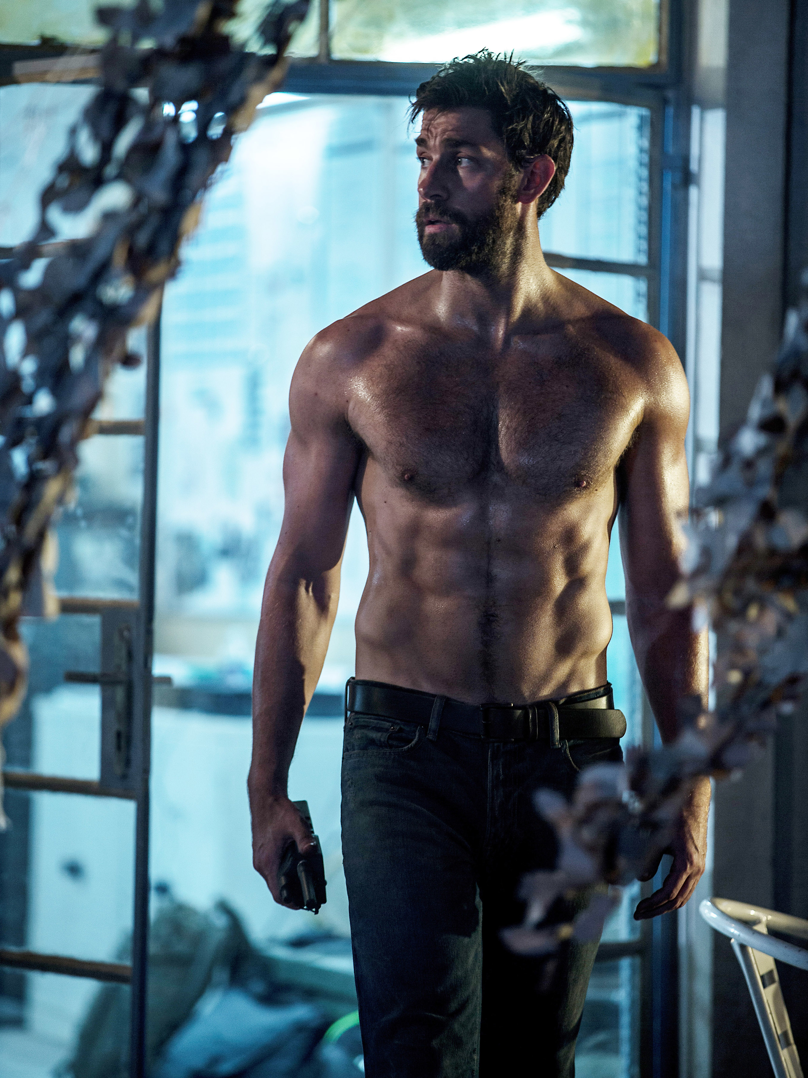 A muscular man with a beard stands shirtless, holding a small object, in an industrial setting with cables and equipment in the background