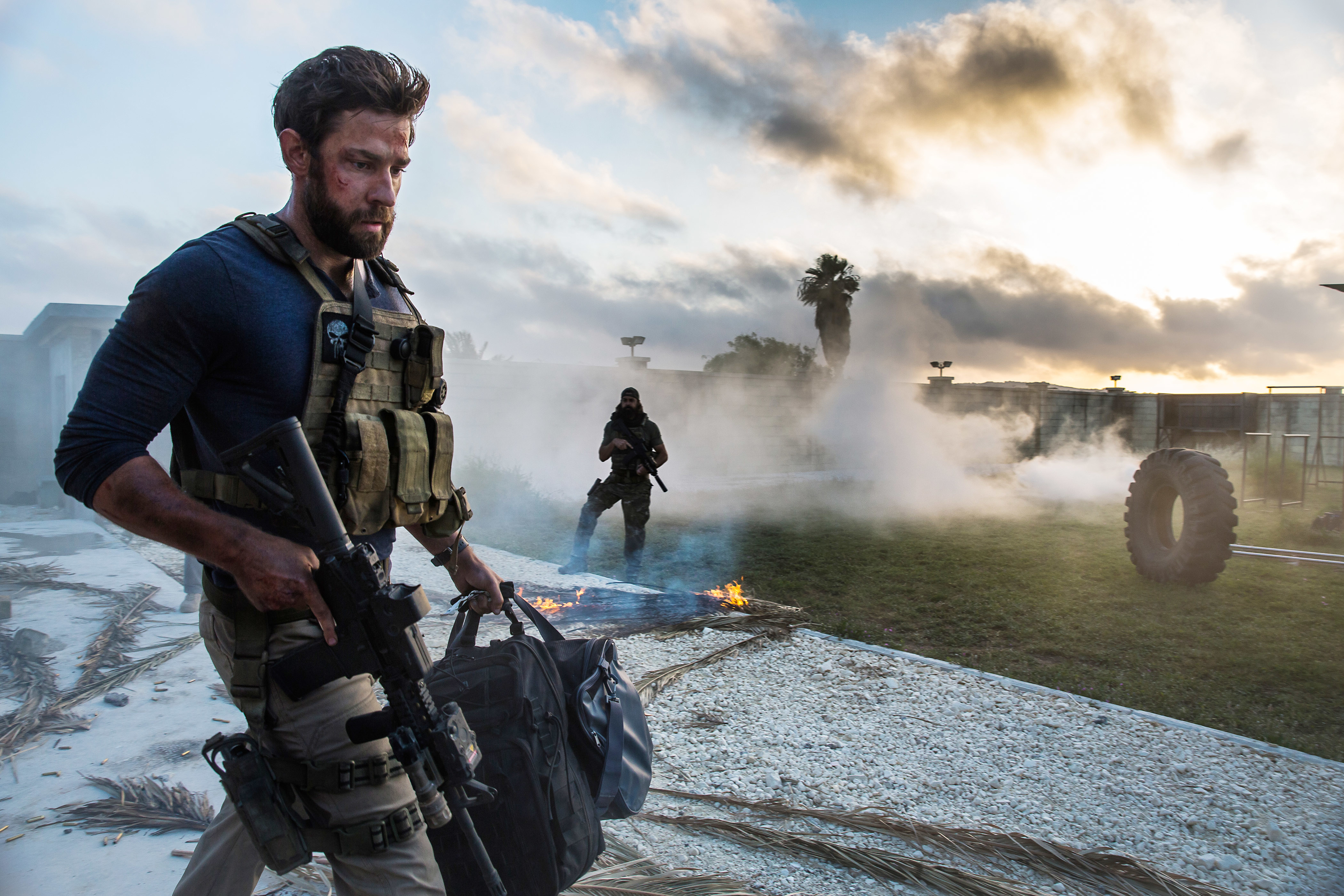 Two people in tactical gear on a smoky outdoor scene, one holding a rifle and bag, with a burning area in the background