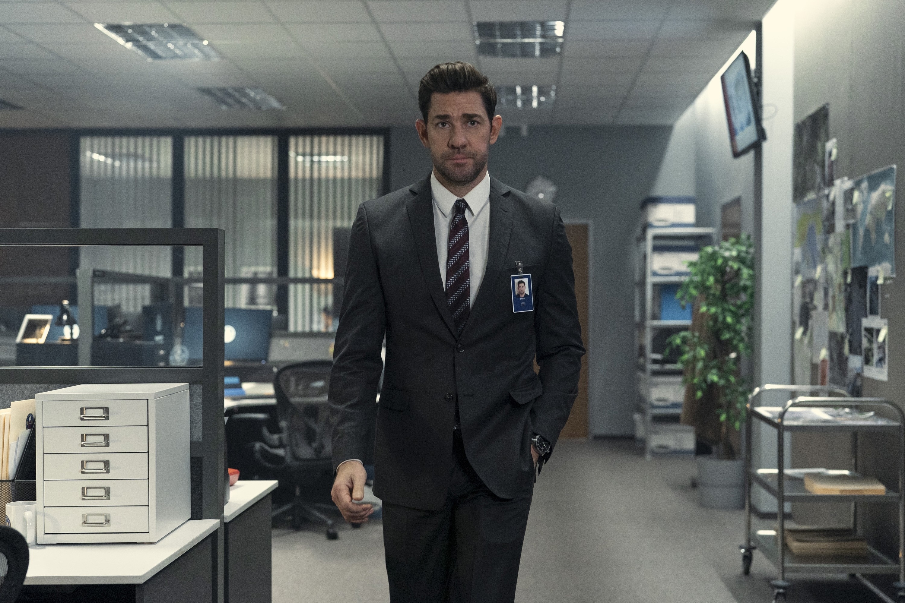 Person in a suit walking through an office setting, wearing a lanyard ID badge. Desks and office equipment are visible in the background