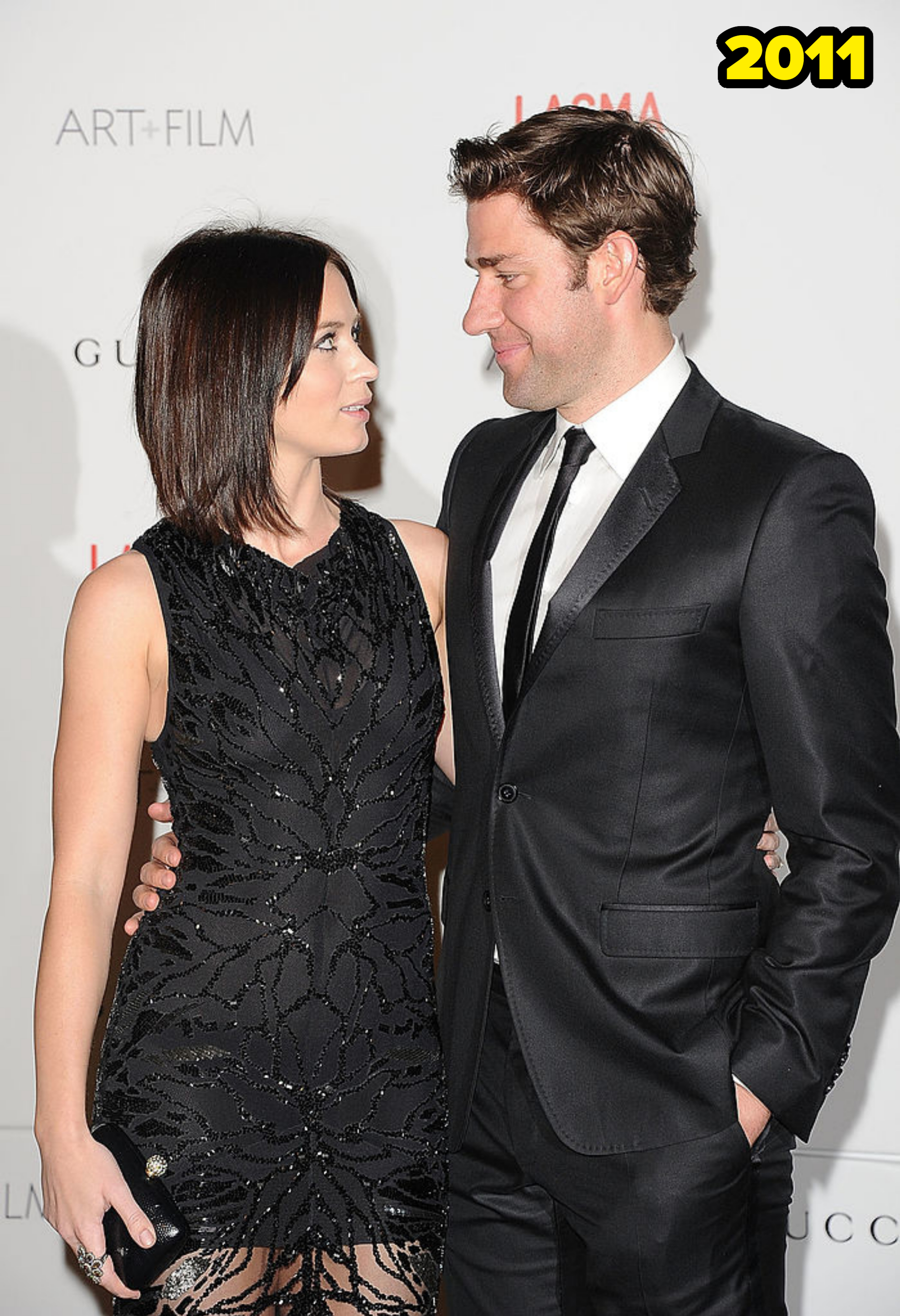 Two people in formal wear, a woman in an elegant patterned dress and a man in a suit, smiling at each other on a red carpet