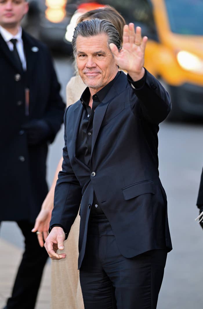Josh Brolin in a sleek suit waves at an event, standing on a street with a yellow taxi in the background