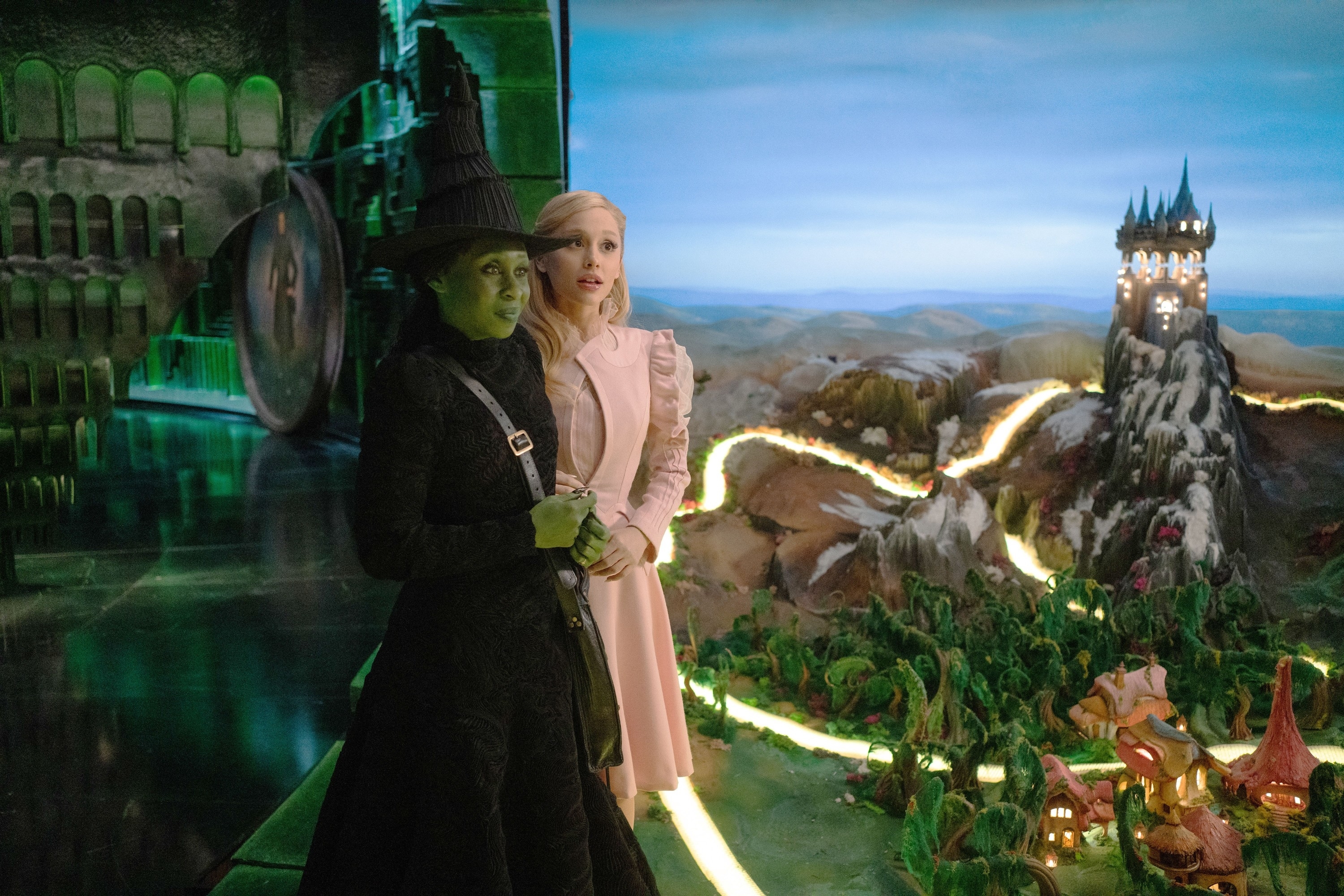 A witch in a black hat and gown stands with a woman in a pink dress, facing a fantasy landscape with a tall tower in the distance