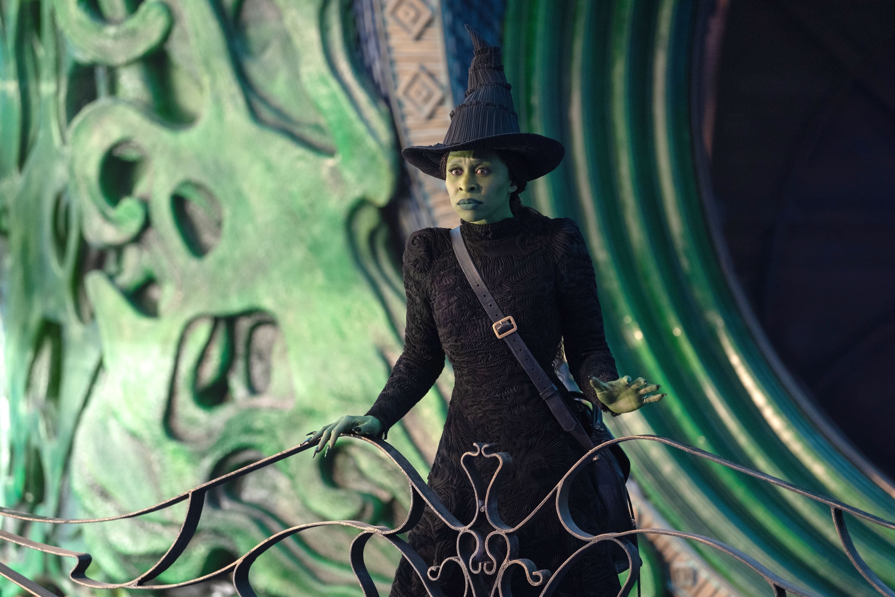 Cynthia Erivo in a theatrical witch costume with green skin and a pointed hat stands on an elaborate set