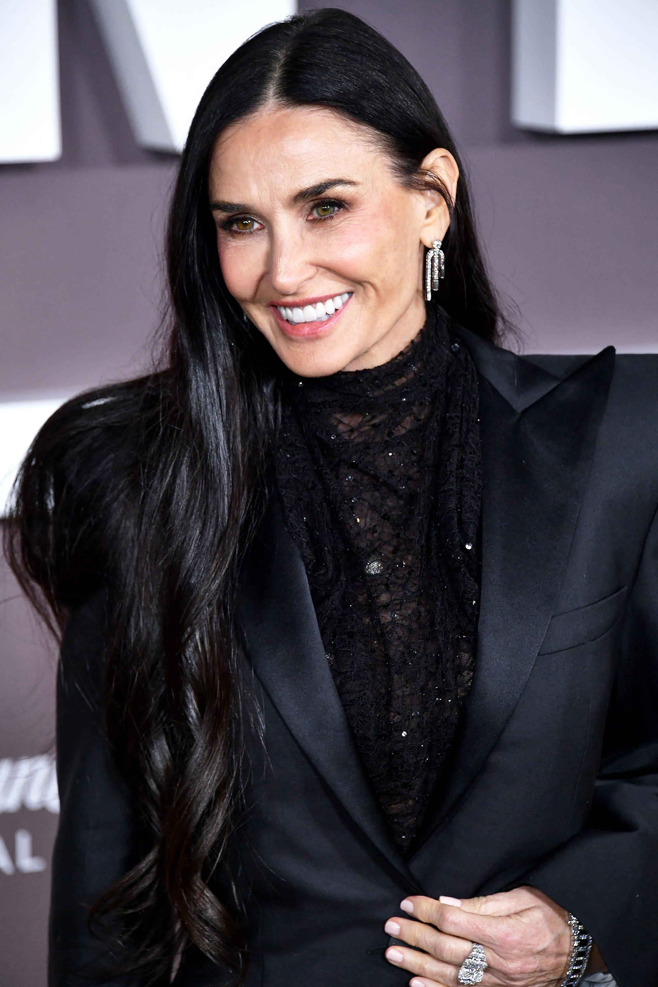 Demi Moore posing on a red carpet in a black lace blouse and a tailored blazer, smiling with long, wavy hair