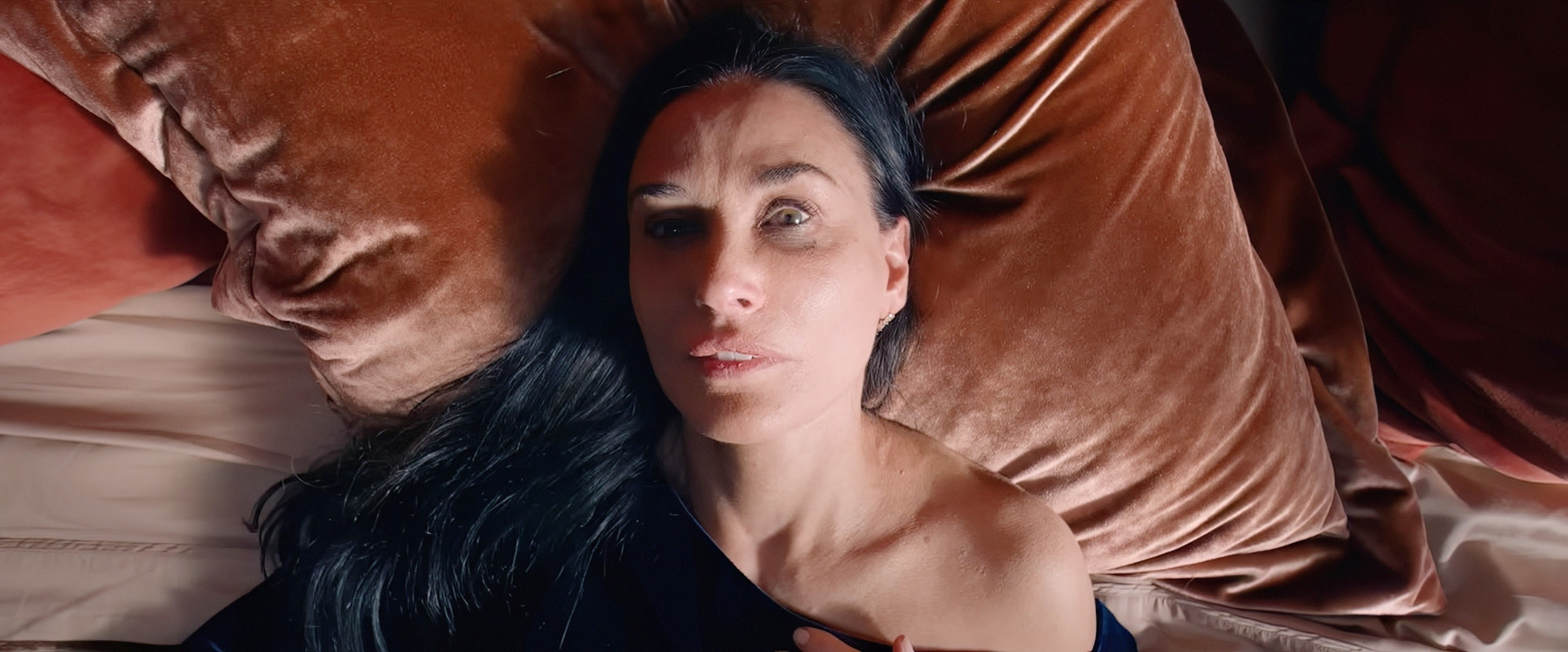 Demi Moore lying on a bed with a thoughtful expression. Dark hair, one shoulder exposed by an off-the-shoulder top. Velvet pillows in the background