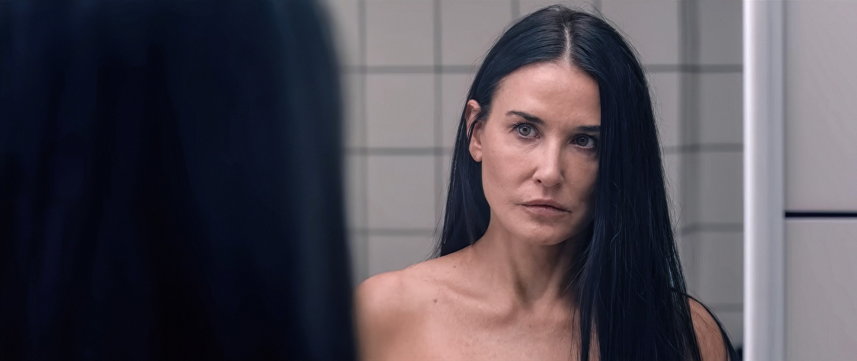 Person with long dark hair looking at themselves in a bathroom mirror, wearing a serious expression