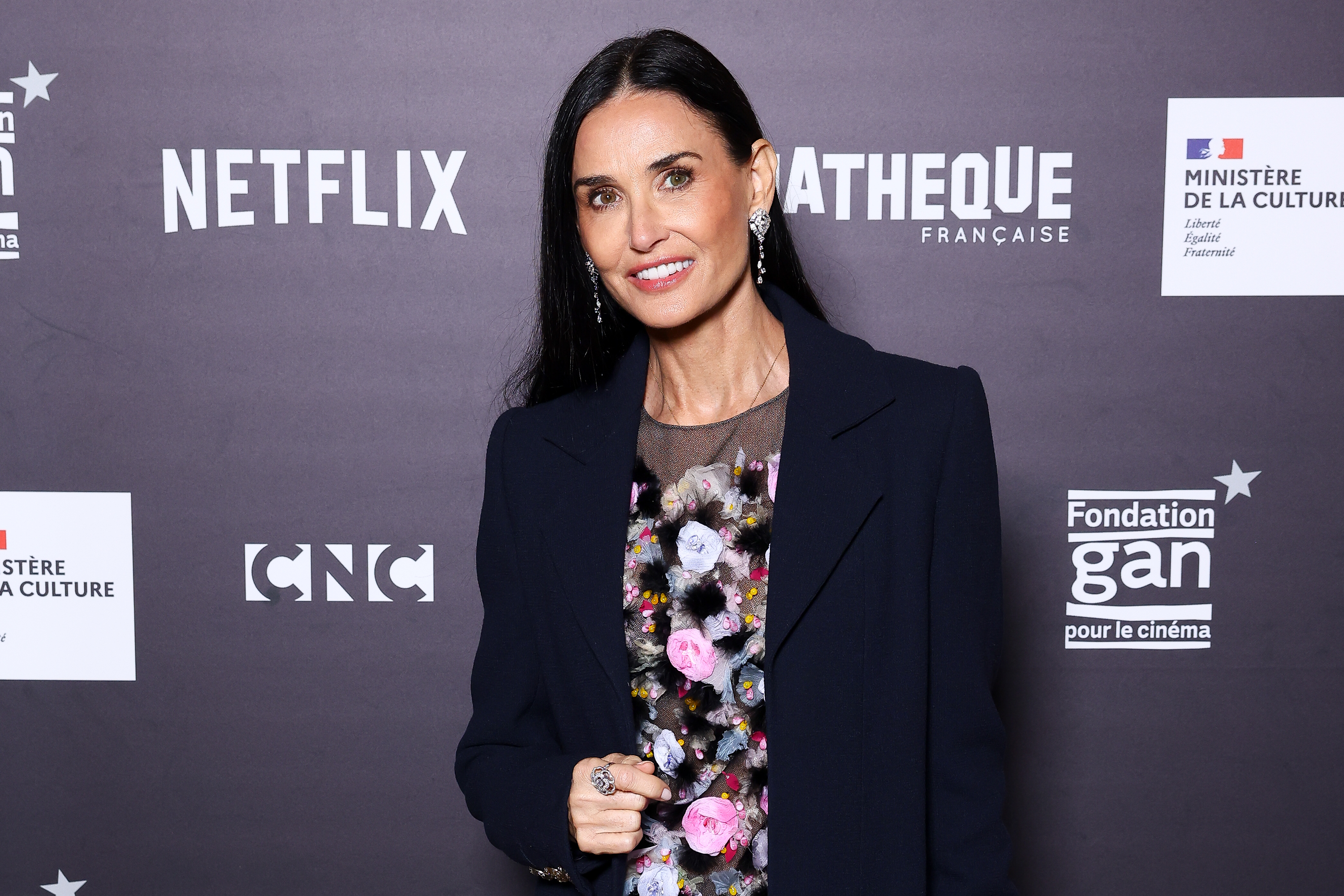 Person posing at a media event, wearing a floral-patterned outfit under a dark blazer, with logos in the background