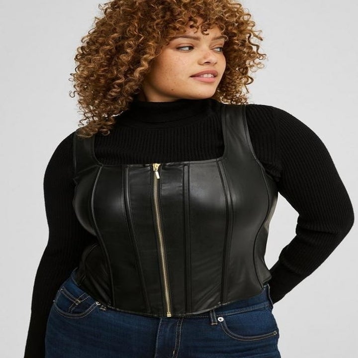 Person wearing a black leather corset over a ribbed turtleneck sweater and jeans, looking to the side