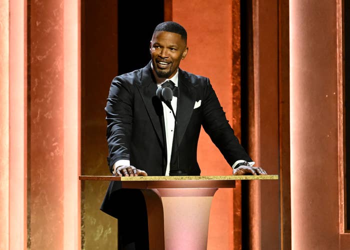 Person in a tuxedo stands at a podium speaking, smiling warmly at an event