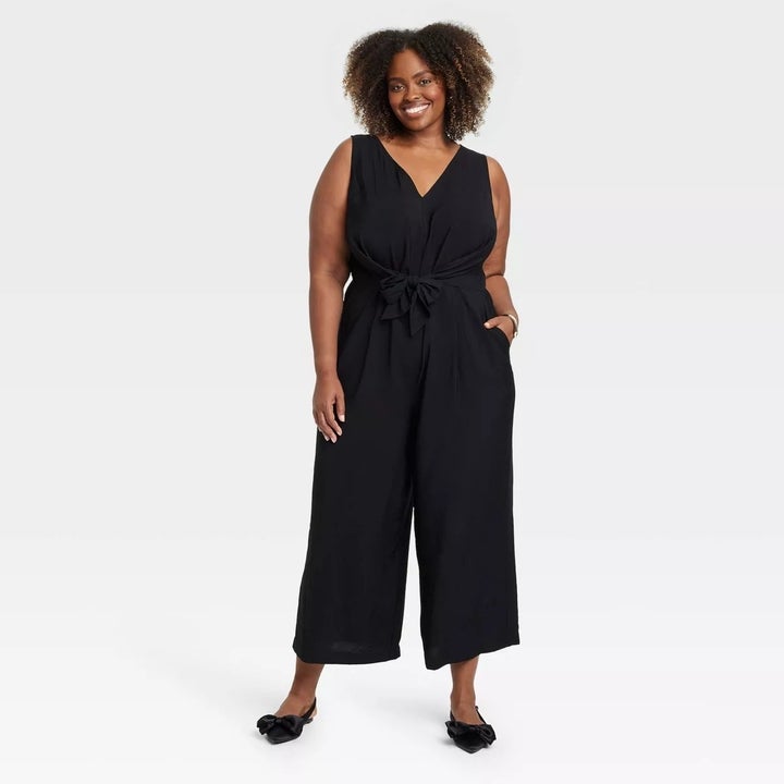 Model wearing a sleeveless black jumpsuit with a waist tie, standing with hands in pockets