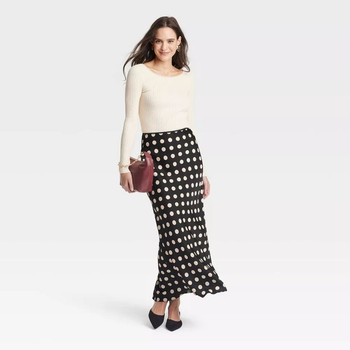 model wearing a fitted long-sleeve top and polka-dot maxi skirt, holding a clutch, standing against a plain background