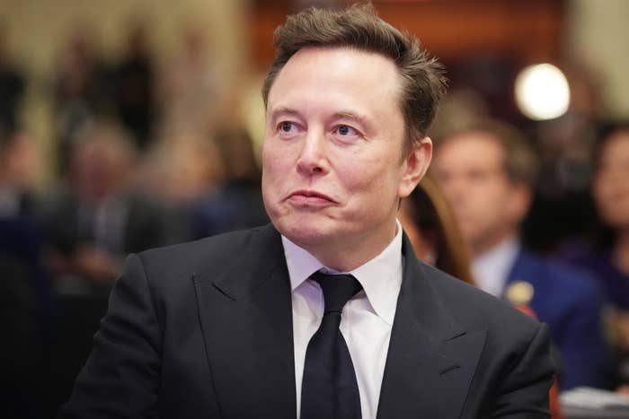 A person in a suit and tie stands in a formal setting, looking to the side