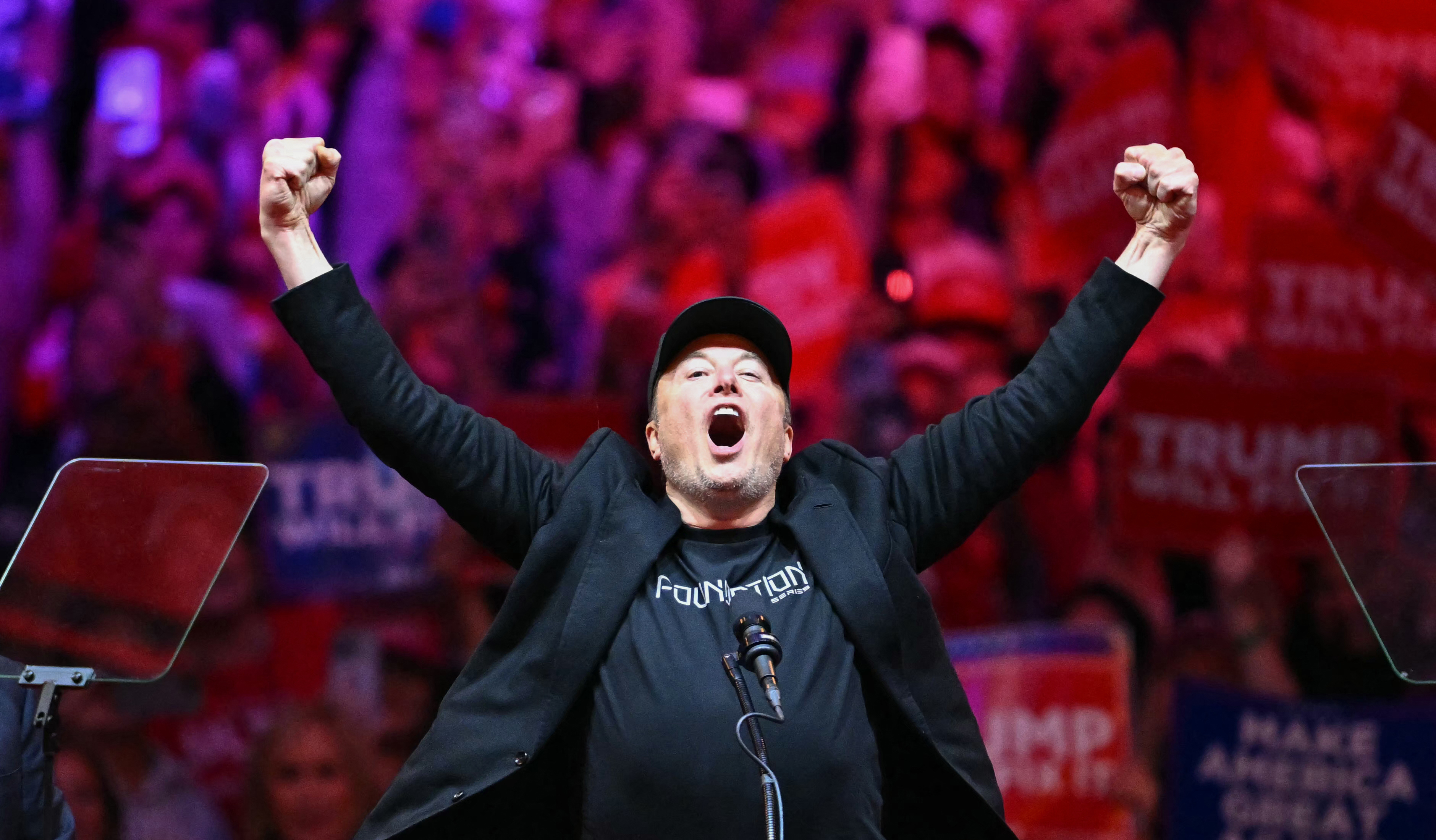 Person in a cap and jacket, arms raised, in front of a cheering crowd with "Trump" signs visible in the background