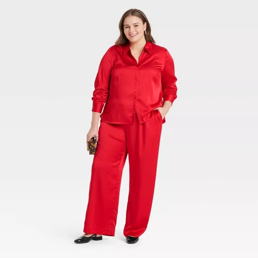A model in a satin blouse and wide-leg pants poses confidently, holding a clutch