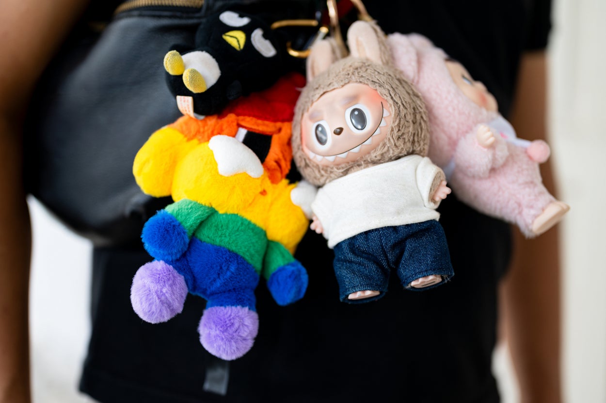 Colorful plush toys attached to a bag, featuring a rainbow bear and a doll in a fuzzy outfit with big eyes