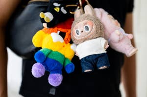 Colorful plush toys attached to a bag, featuring a rainbow bear and a doll in a fuzzy outfit with big eyes