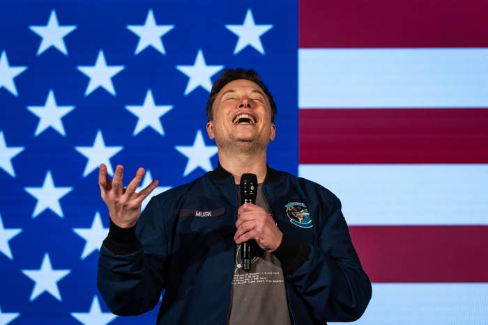 Person laughing while holding a microphone, standing in front of a U.S. flag backdrop. Wearing a jacket with a patch