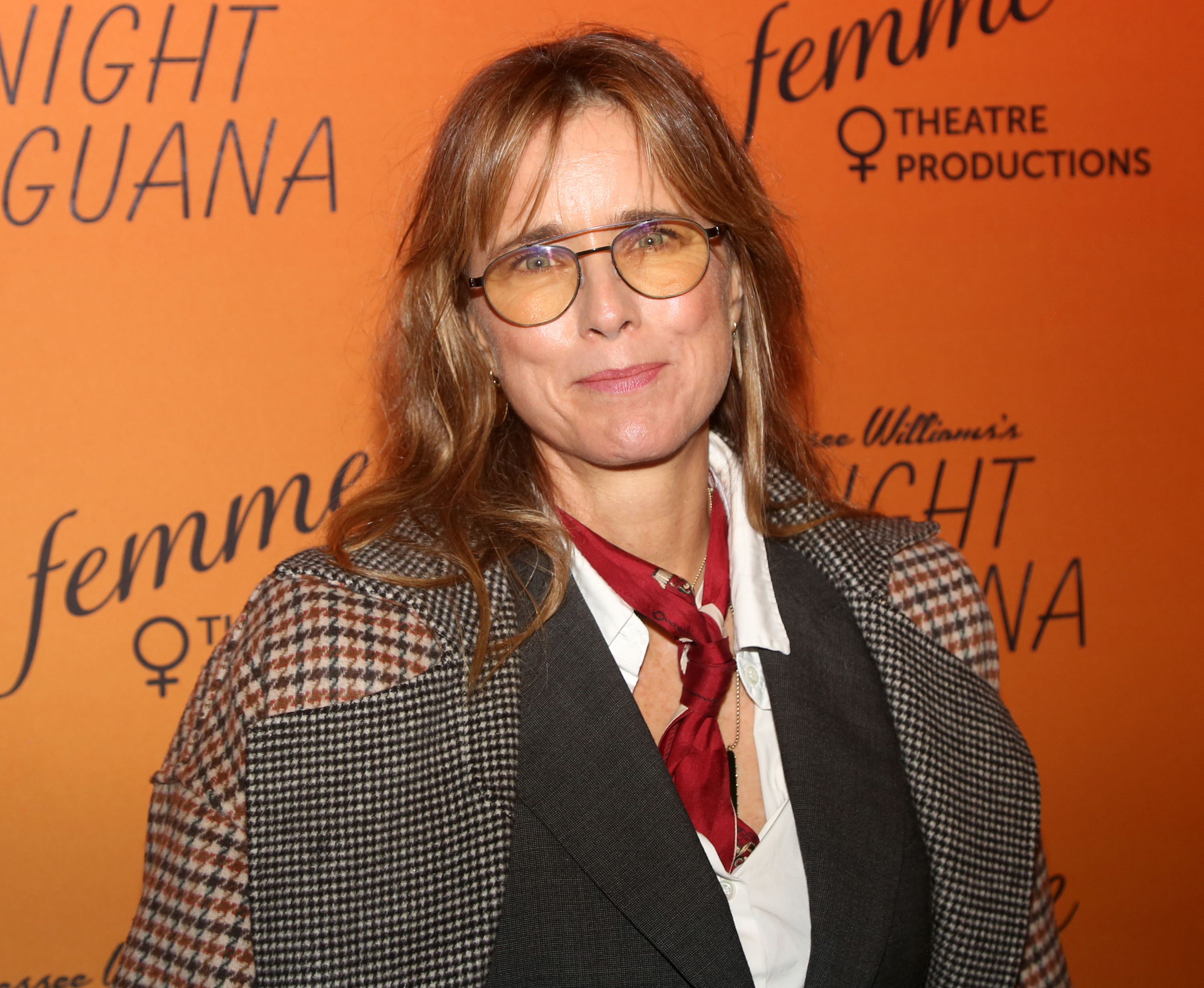 Person on the red carpet wearing a houndstooth blazer, white shirt, and red scarf at a theatre production event