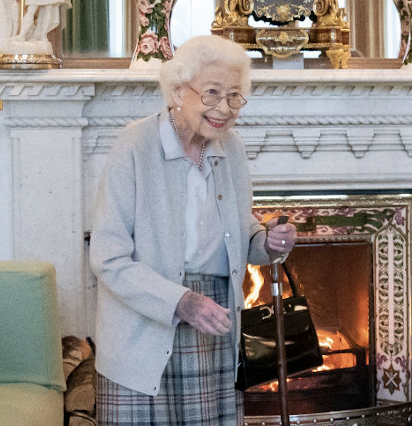 An elderly woman with glasses and a plaid skirt stands smiling, holding a cane, in a cozy room with a fireplace in the background