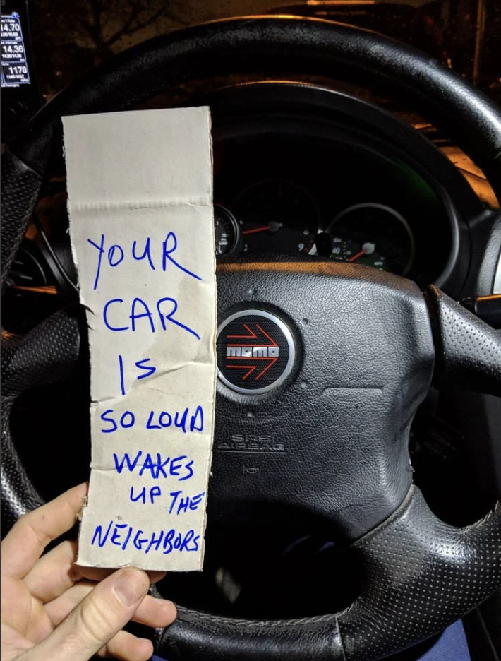 Hand holding sign reading, &quot;YOUR CAR is SO LOUD WAKES UP THE NEIGHBORS,&quot; inside a car with hands on the wheel