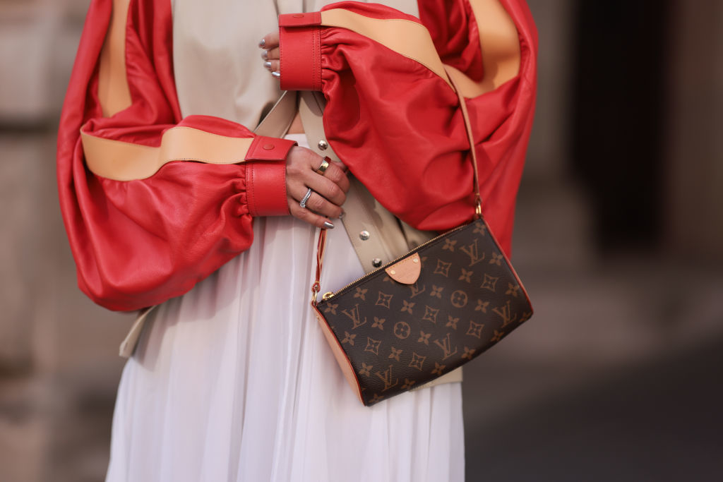 Person wearing a voluminous red and beige top with a brown designer handbag. Hands are adorned with rings over a light-colored skirt