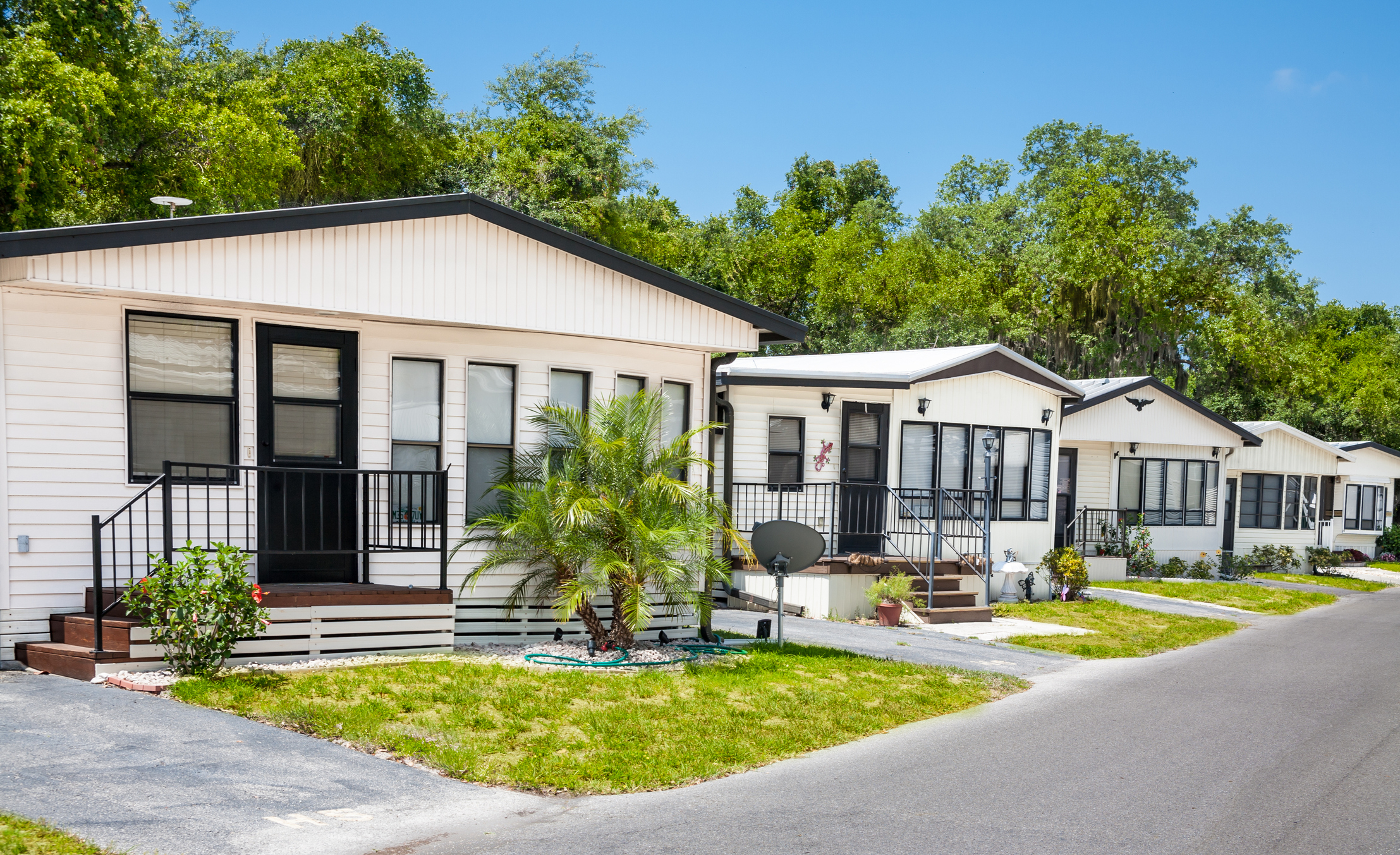 Row of mobile homes on a sunny day, surrounded by greenery, with a street in the foreground