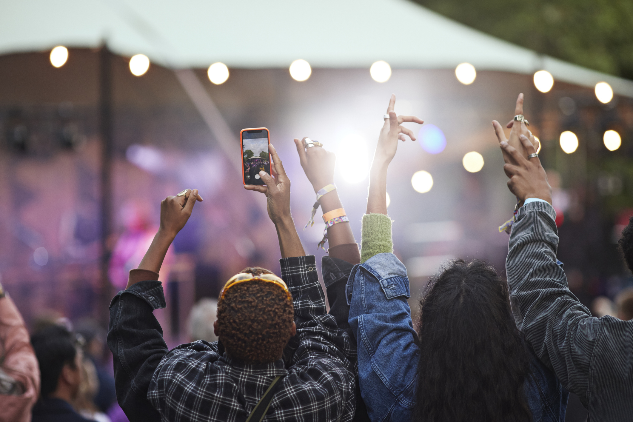 Audience at an outdoor concert raising hands and phones, engaging with the performance under string lights