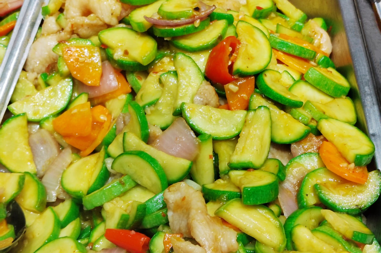 Stir-fried dish with sliced zucchini, bell peppers, and chicken, displayed in a buffet tray with a serving spoon nearby