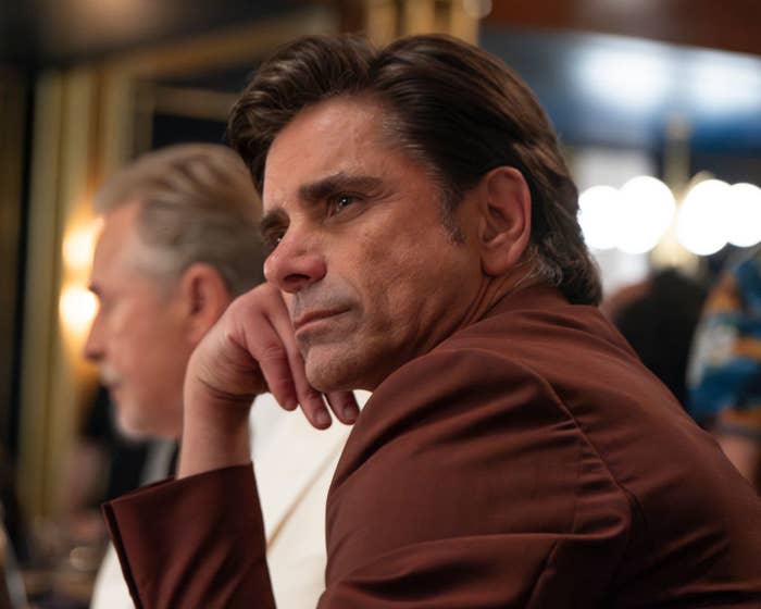 A man in a formal setting, wearing a brown jacket, sits thoughtfully with his chin resting on his hand. Another man is blurred in the background