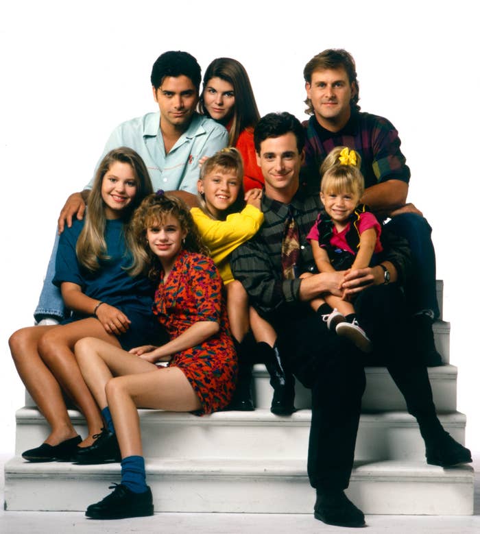 Group photo of cast members from a TV family sitcom, sitting and standing on white stairs, dressed in casual '90s style attire
