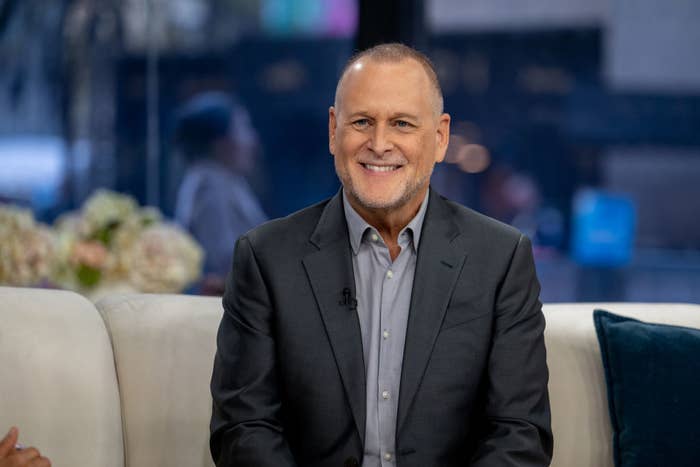 Man in a suit sits smiling on a couch during a talk show interview