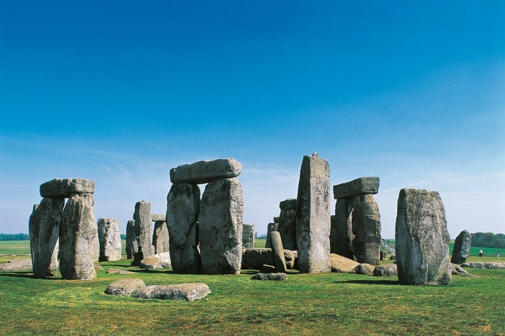 Stonehenge under a clear sky with scattered clouds, surrounded by green grass and distant visitors exploring the historic site