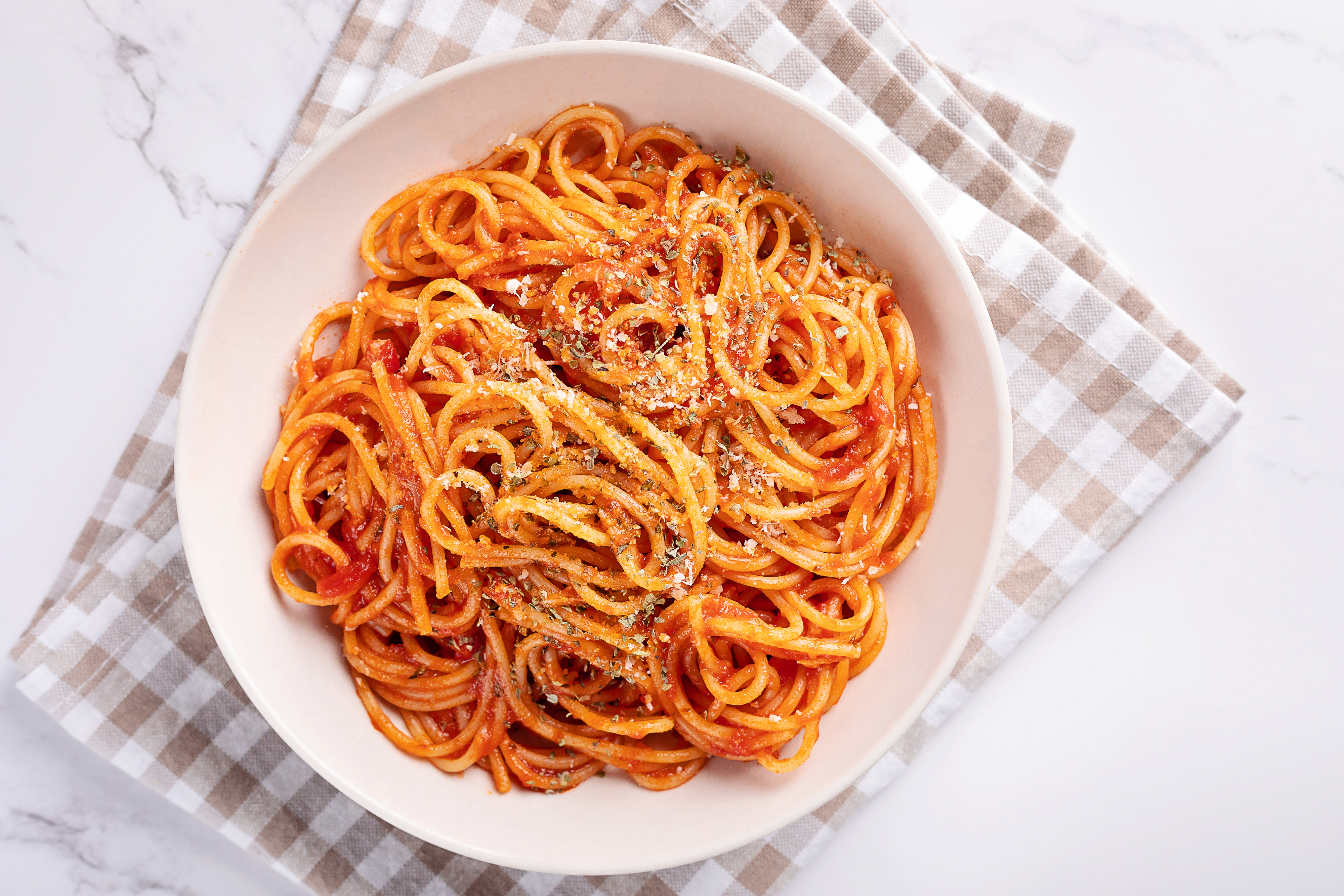 Bowl of spaghetti with tomato sauce and sprinkled cheese on a checkered cloth and marble counter.