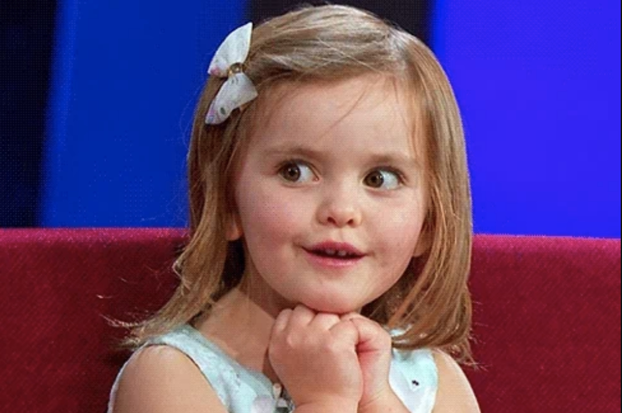 Young child with hands under chin, wearing a light dress and a hair clip, looking surprised or excited