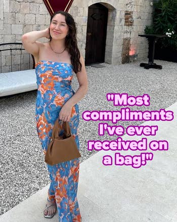 Person in a floral-patterned dress poses outdoors, holding a brown handbag. Stone wall and decorative banner in the background