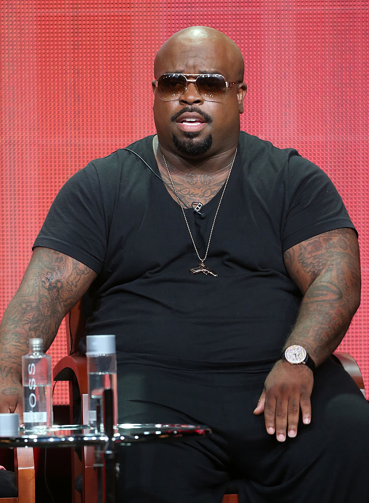 Individual sitting on a panel, wearing a V-neck T-shirt and sunglasses. There is a water bottle on a table next to them