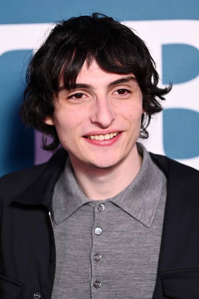 A person with short, dark hair smiles at the camera. They wear a gray collared shirt under a dark jacket