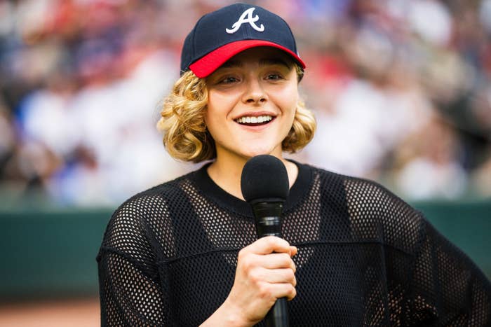 A person wearing a baseball cap and mesh top holds a microphone, smiling at an outdoor event