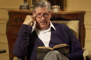 A person wearing glasses and a cardigan is sitting in a chair, reading a book and looking forward thoughtfully