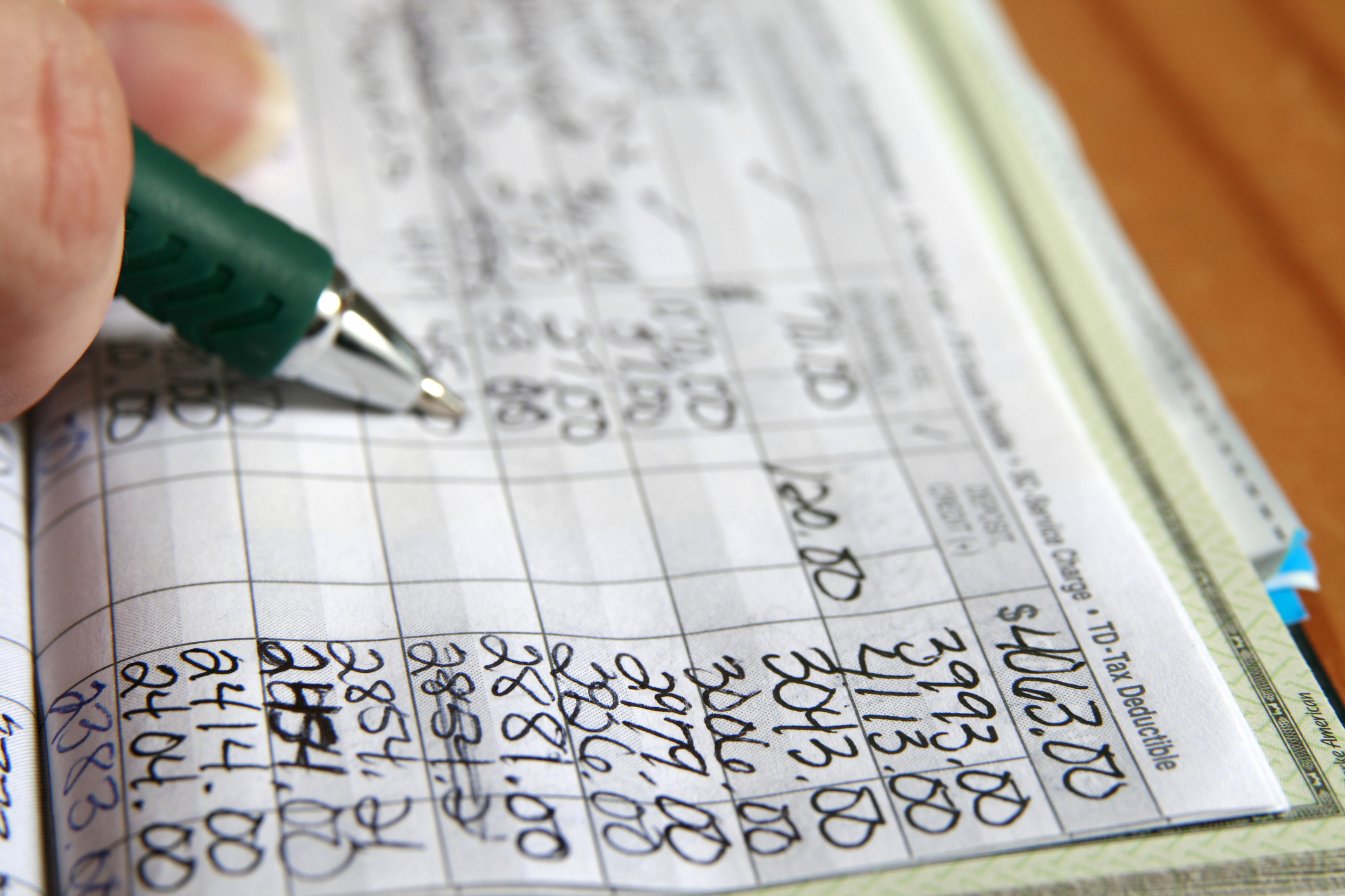 Hand writing in a checkbook register with a pen, showing various transactions and balances