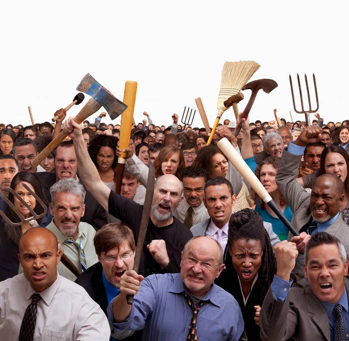 A diverse crowd of people in office wear holding tools like pitchforks and brooms, expressing anger and protest