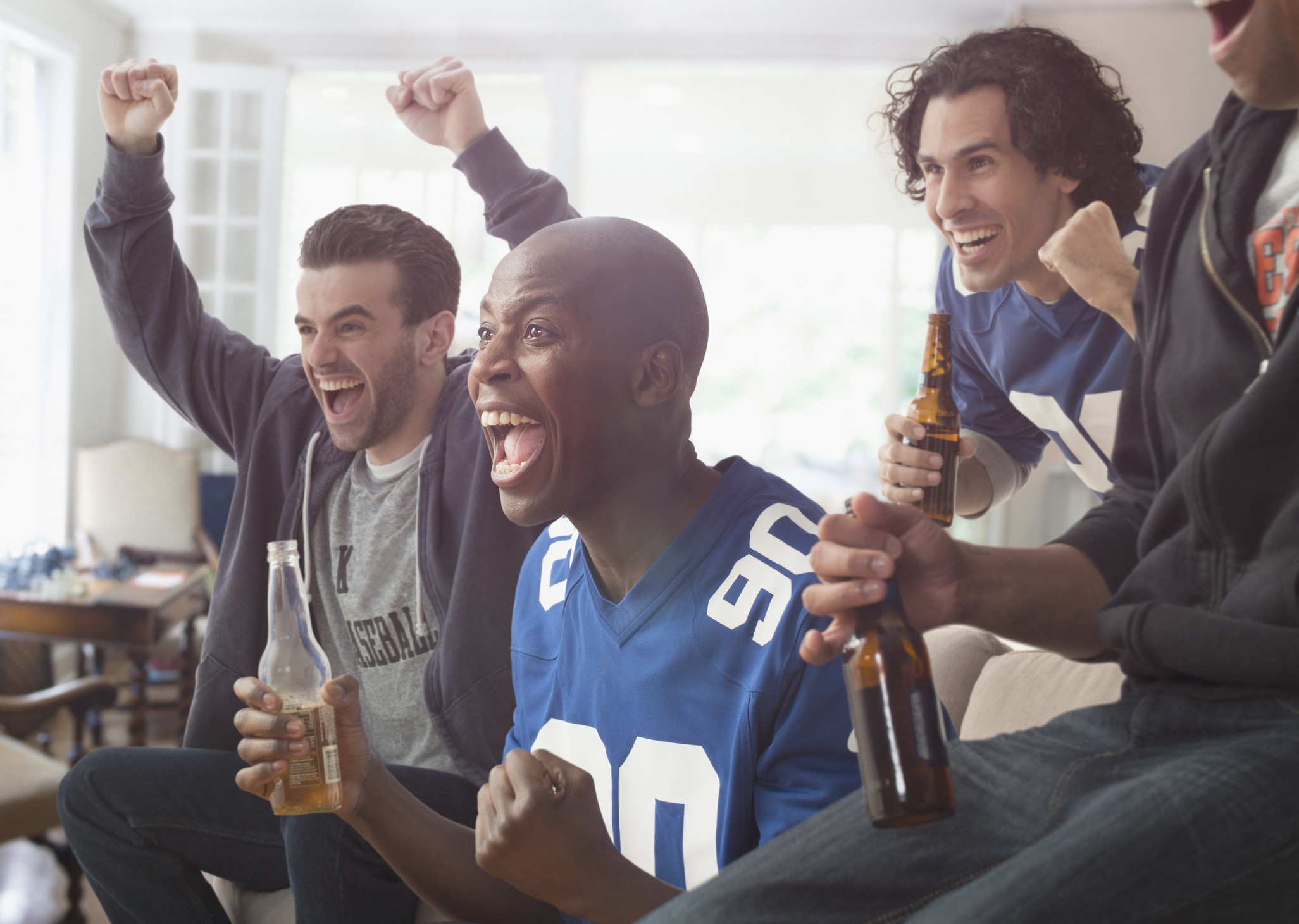Four excited individuals indoors celebrate with drinks, wearing casual sports jerseys, showing enthusiasm and joy