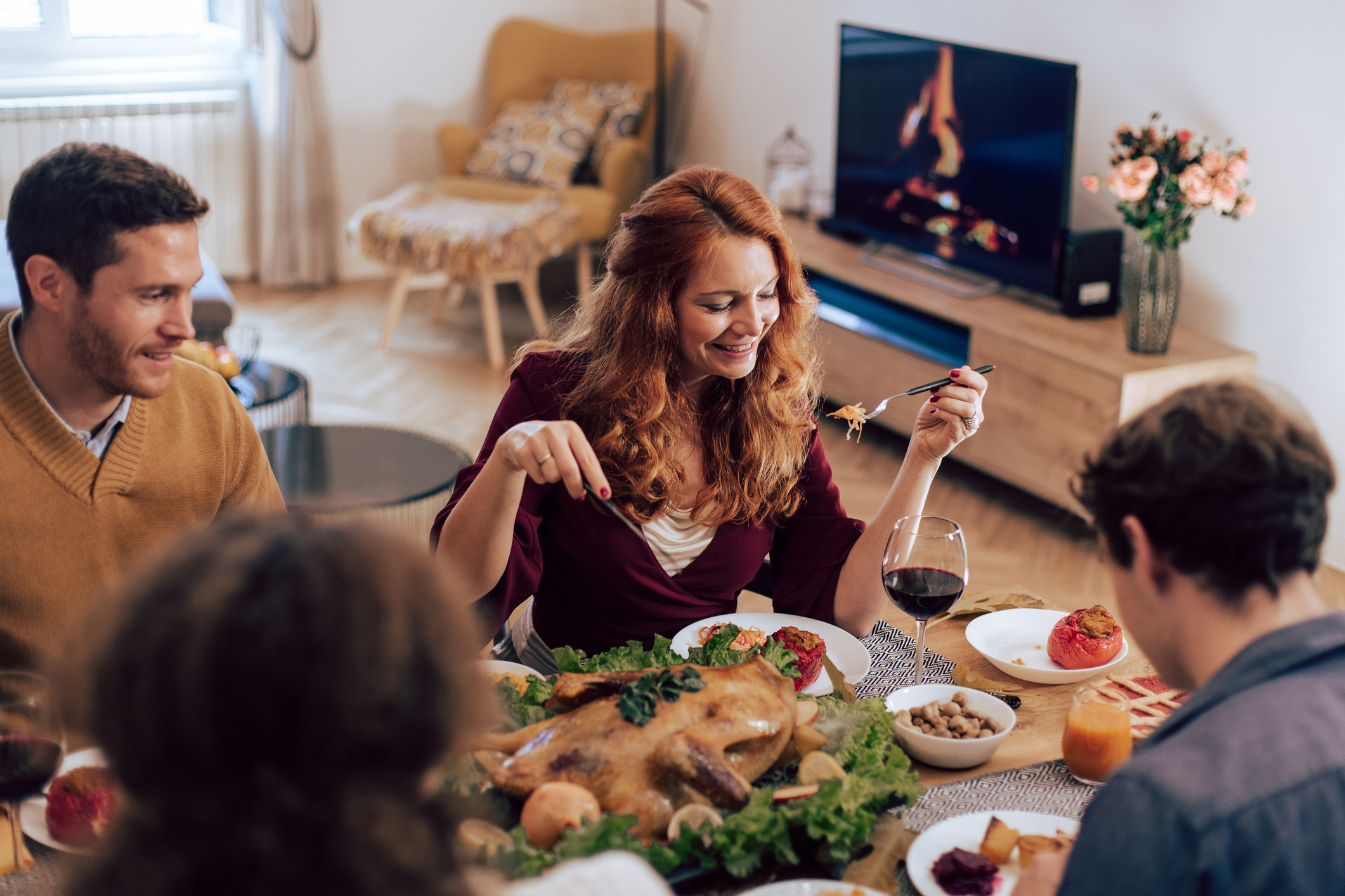 People gathered around a dining table enjoying a meal, with a whole roast pig centerpiece and a woman holding a glass of wine, smiling