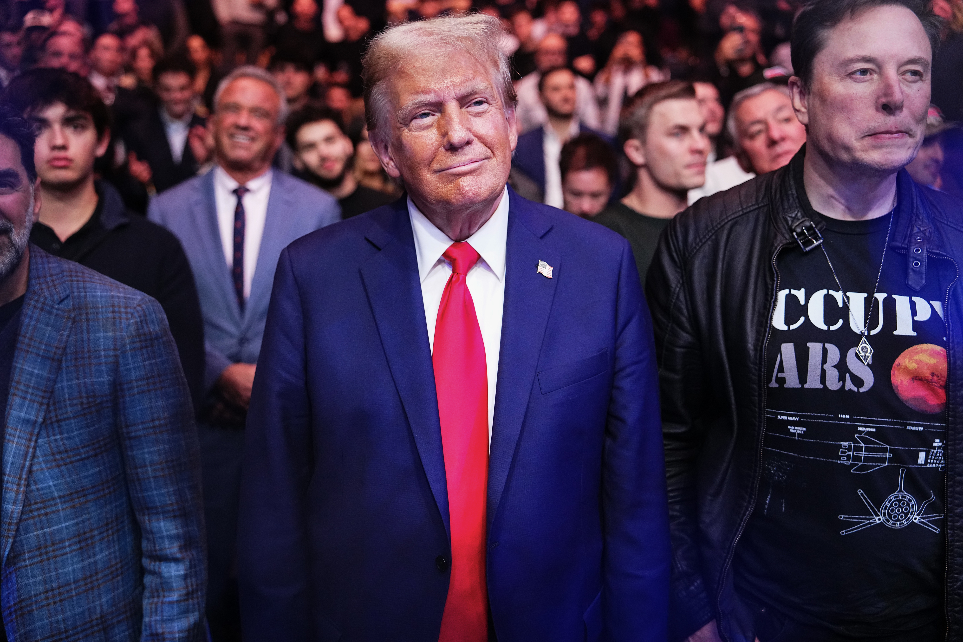 A man in a suit and tie stands among a crowd, with a focus on his attire and expression